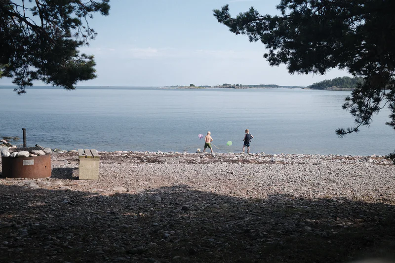 Two people walk along a rocky shoreline by a calm lake, framed by overhanging pine branches.