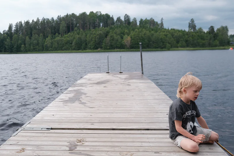 Young boy sits on the edge of a wooden dock over a lake, with forested shoreline in the background.