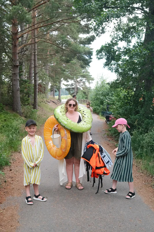 Smiling adults and children walk along a wooded trail holding large inflatable pool toys.