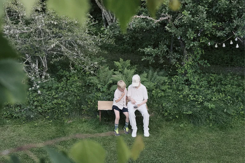 Two people in caps sit on a small bench in a green garden with trees and plants around them.