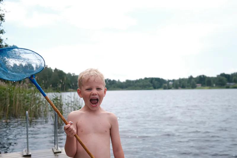 Smiling shirtless child on a lakeside boat holds a blue net pole as water and trees fill the background.