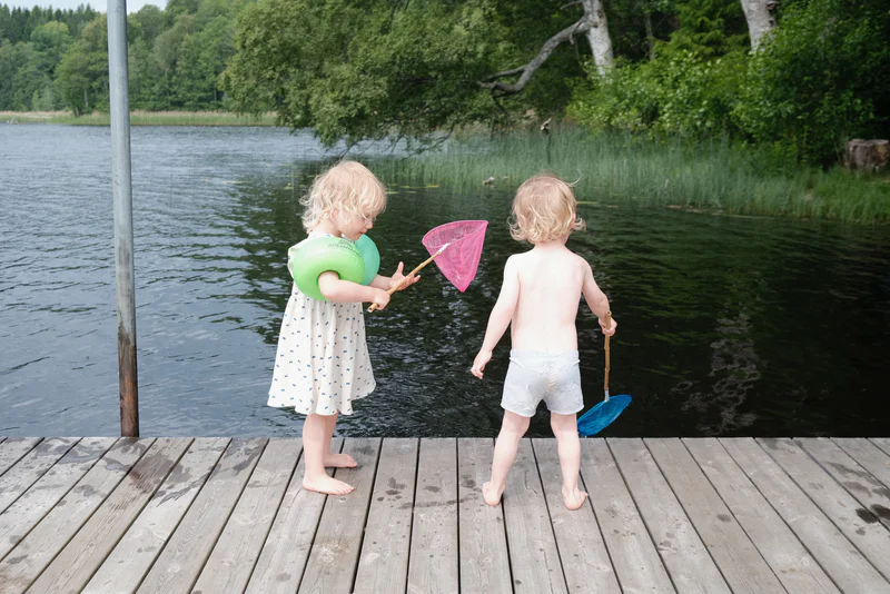 Two toddlers stand on a lakeside dock holding bath toys and fishing nets, with trees and water behind them.
