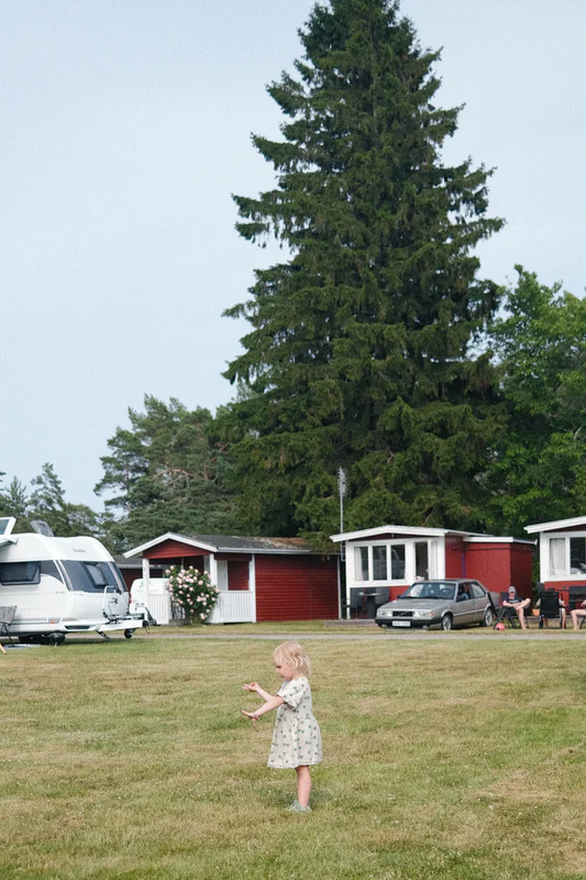Child in a white dress walks on grass in front of an airplane and red-and-white houses.