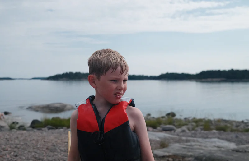 Young boy in a red-and-black life jacket standing by a calm lake with rocky shore in the background.