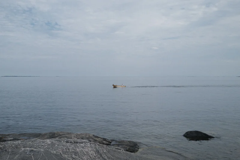 A small boat floats on calm gray water near rocky shore under an overcast sky.